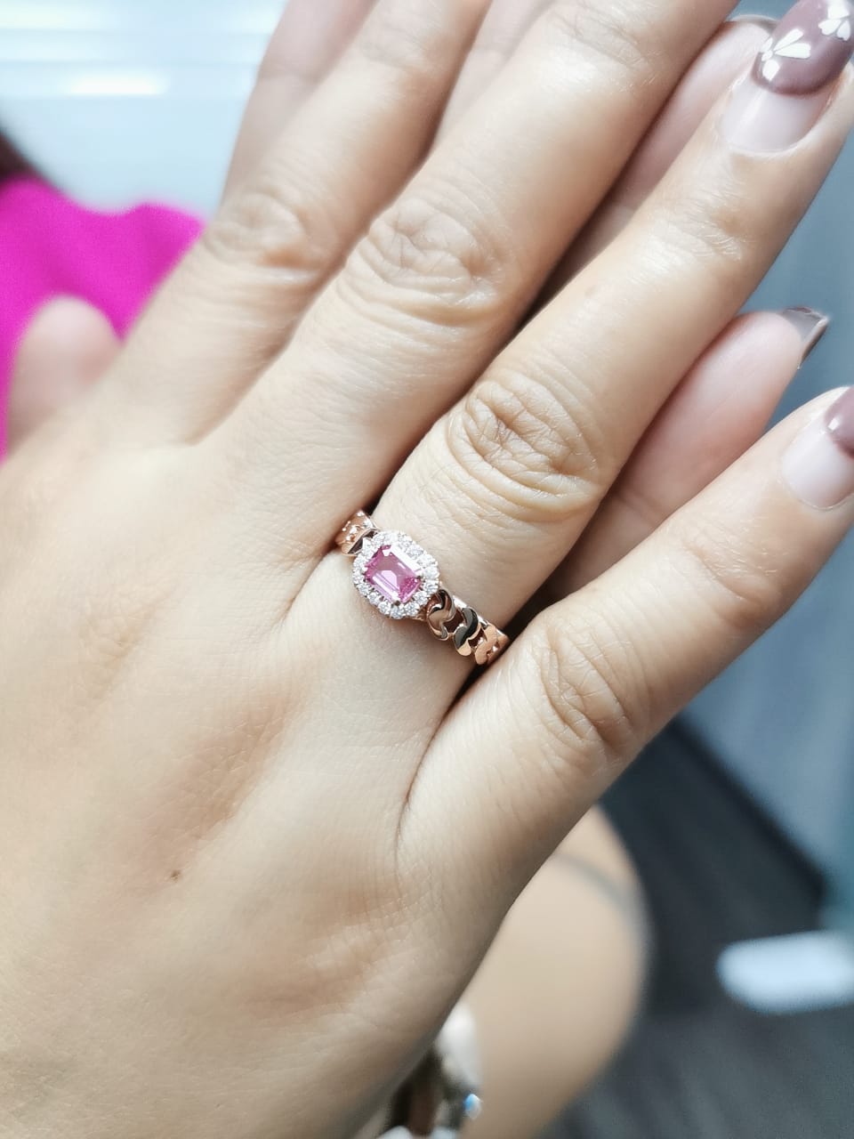 Elegant 18K gold pink sapphire and diamond halo ring with a 0.50-carat emerald-cut pink sapphire, surrounded by 0.14-carat round diamonds and a chain-link band design.