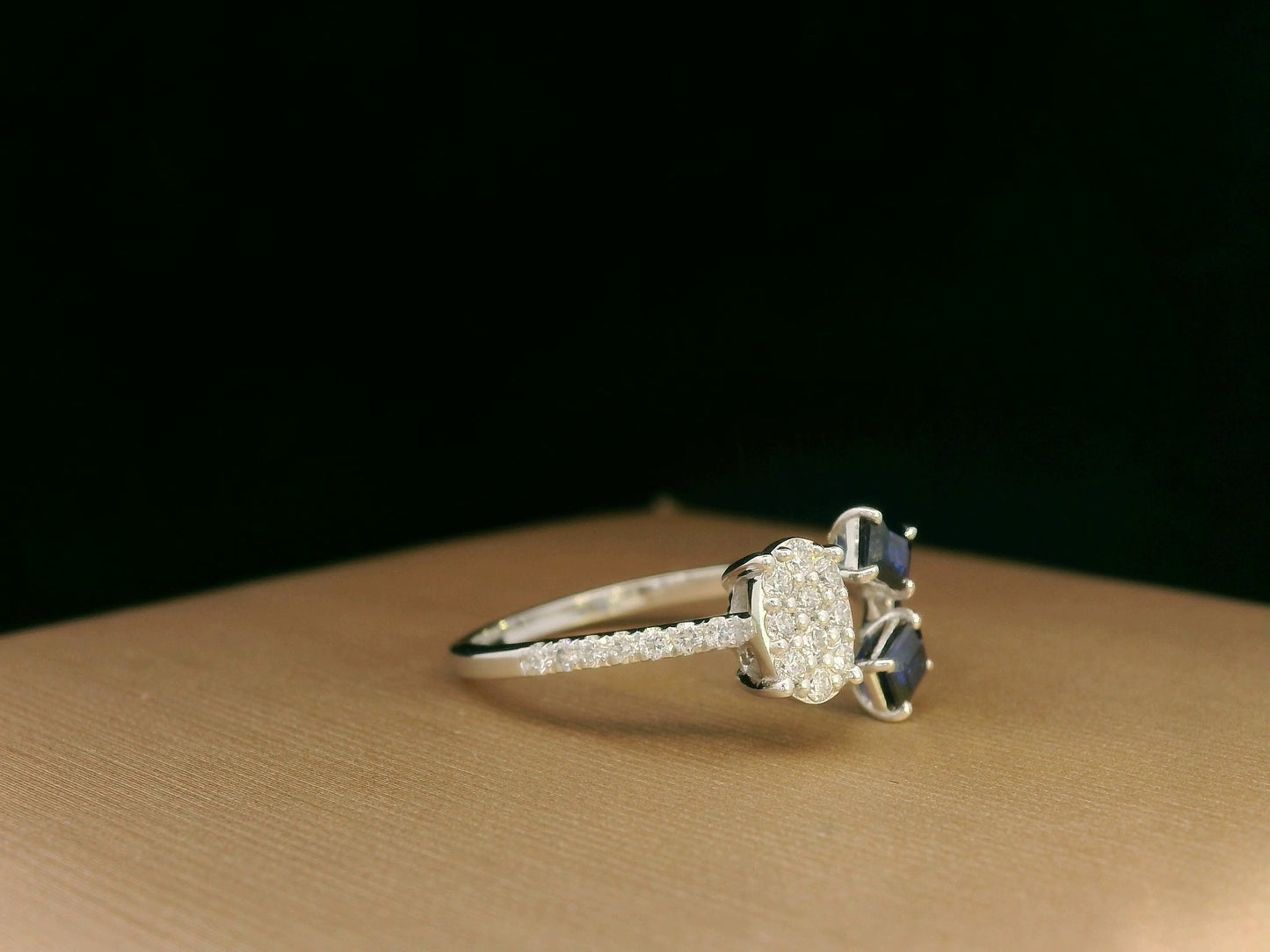 18K white gold open-shank ring featuring an oval diamond cluster on one side and two emerald-cut blue sapphires on the other, accented with pavé diamonds along the band.