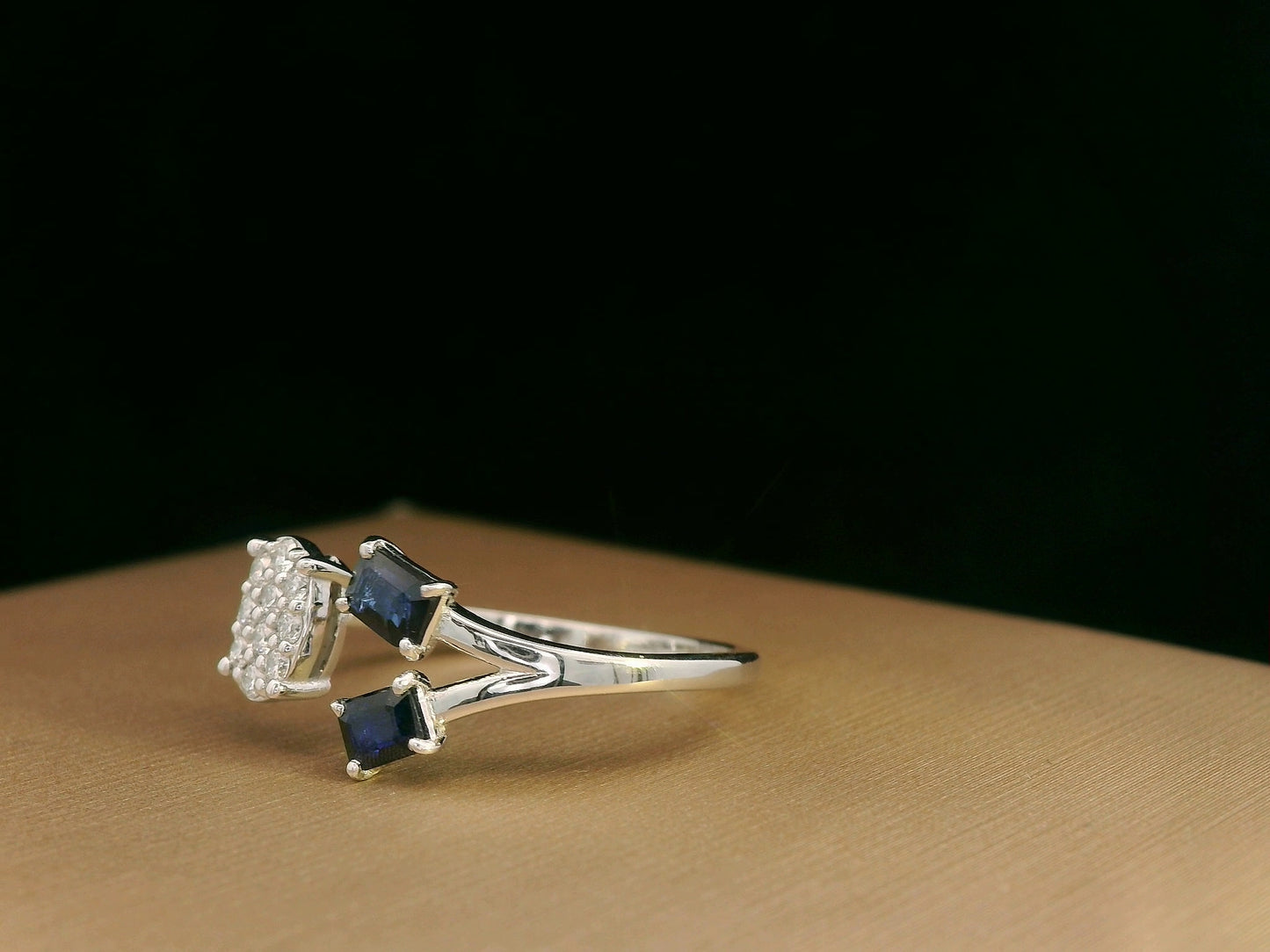 18K white gold open-shank ring featuring an oval diamond cluster on one side and two emerald-cut blue sapphires on the other, accented with pavé diamonds along the band.