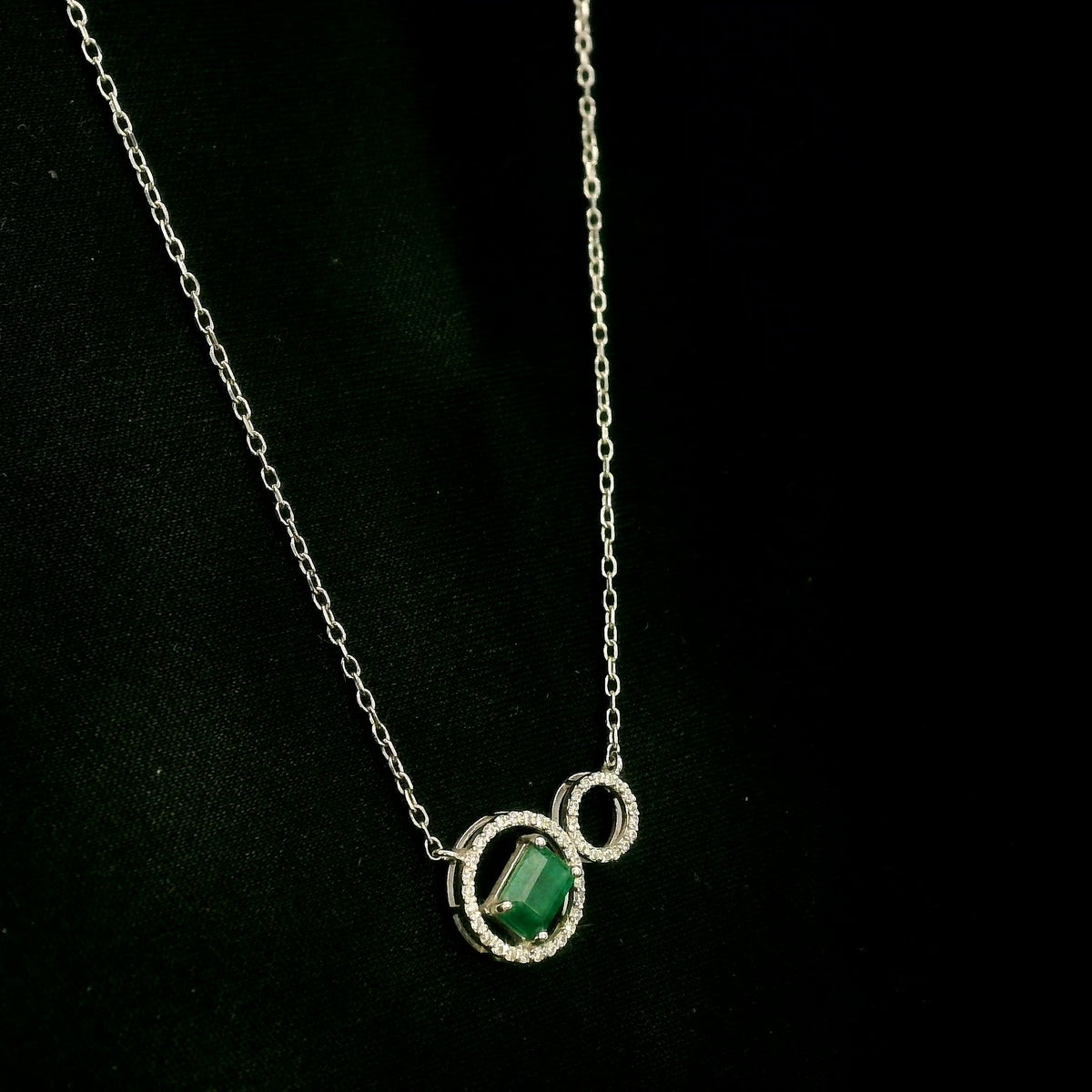 Emerald and diamond necklace in 18K white gold featuring a rectangular emerald-cut green gemstone inside a diamond circle, connected to a smaller diamond-set circle.