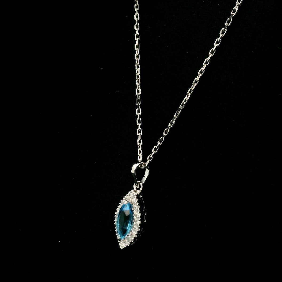 Blue topaz and diamond halo pendant in 18K white gold featuring a 0.70 carat oval blue topaz surrounded by 0.15 carat round diamonds in an elongated marquise-shaped setting, shown with a chain for display.