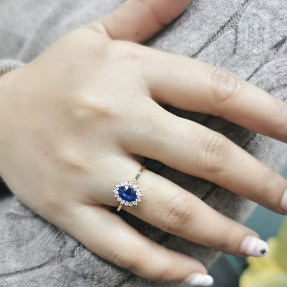 Oval Blue Sapphire and diamond halo ring in 18K gold, featuring a 1.02ct royal blue gemstone surrounded by 0.19ct round diamonds. Elegant fine jewelry inspired by Princess Diana’s iconic ring.