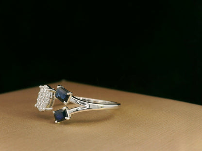 18K white gold open-shank ring featuring an oval diamond cluster on one side and two emerald-cut blue sapphires on the other, accented with pavé diamonds along the band.