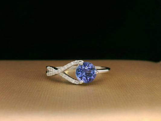 18K white gold ring featuring a 0.72ct round-cut tanzanite with 0.10ct criss-cross diamond accent band on one side and a smooth polished band on the other.