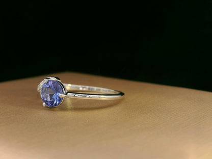 18K white gold ring featuring a 0.72ct round-cut tanzanite with 0.10ct criss-cross diamond accent band on one side and a smooth polished band on the other.