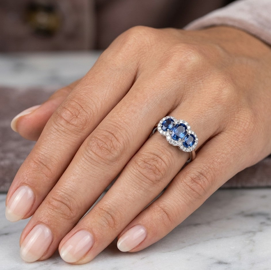 Three stone blue sapphire ring in 18K white gold featuring oval sapphires surrounded by individual diamond halos.