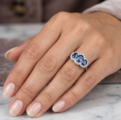 Three stone blue sapphire ring in 18K white gold featuring oval sapphires surrounded by individual diamond halos.