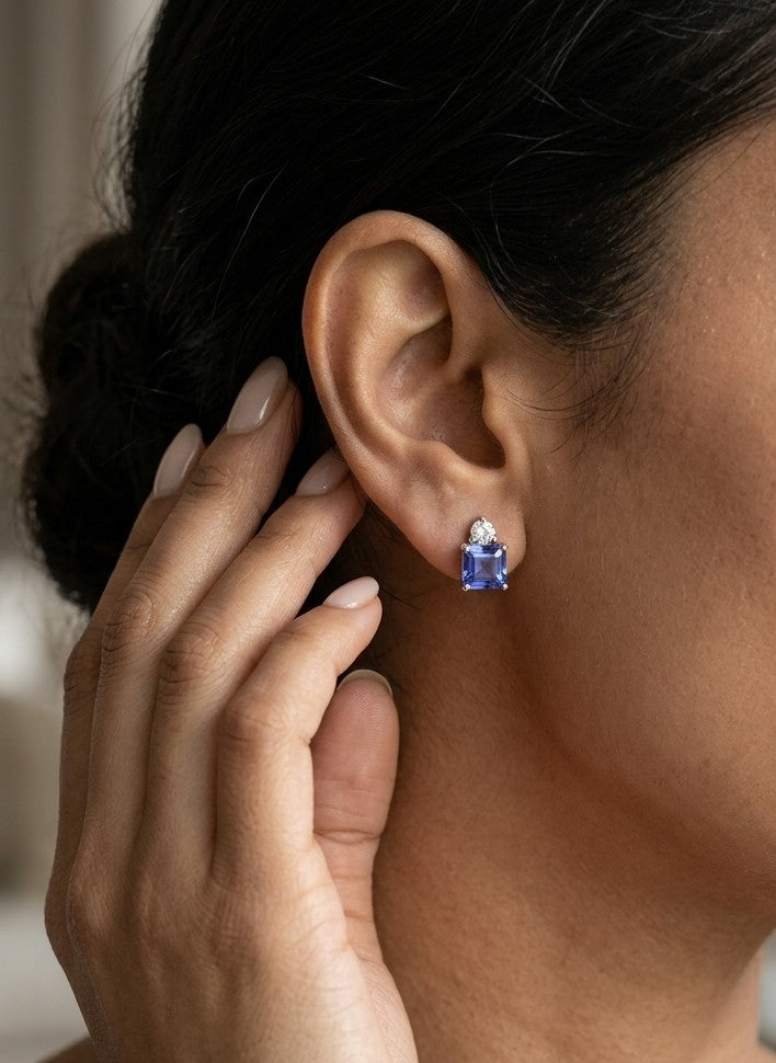Pair of 18K white gold tanzanite and diamond stud earrings featuring square emerald-cut blue-violet tanzanites with a round diamond accent above each gemstone.