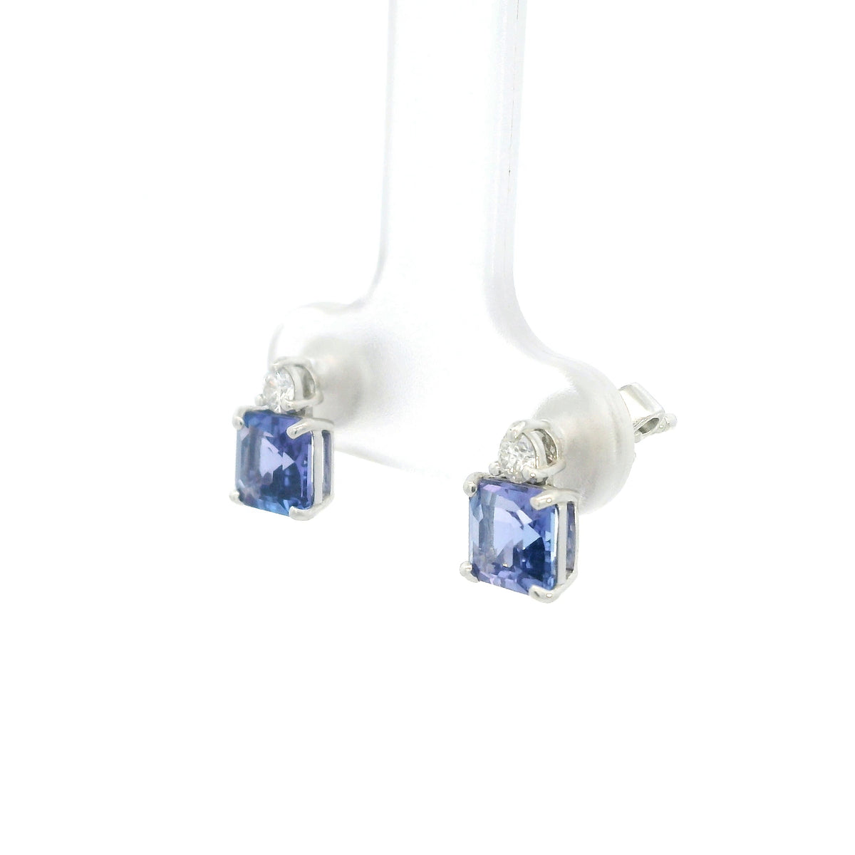 Pair of 18K white gold tanzanite and diamond stud earrings featuring square emerald-cut blue-violet tanzanites with a round diamond accent above each gemstone.