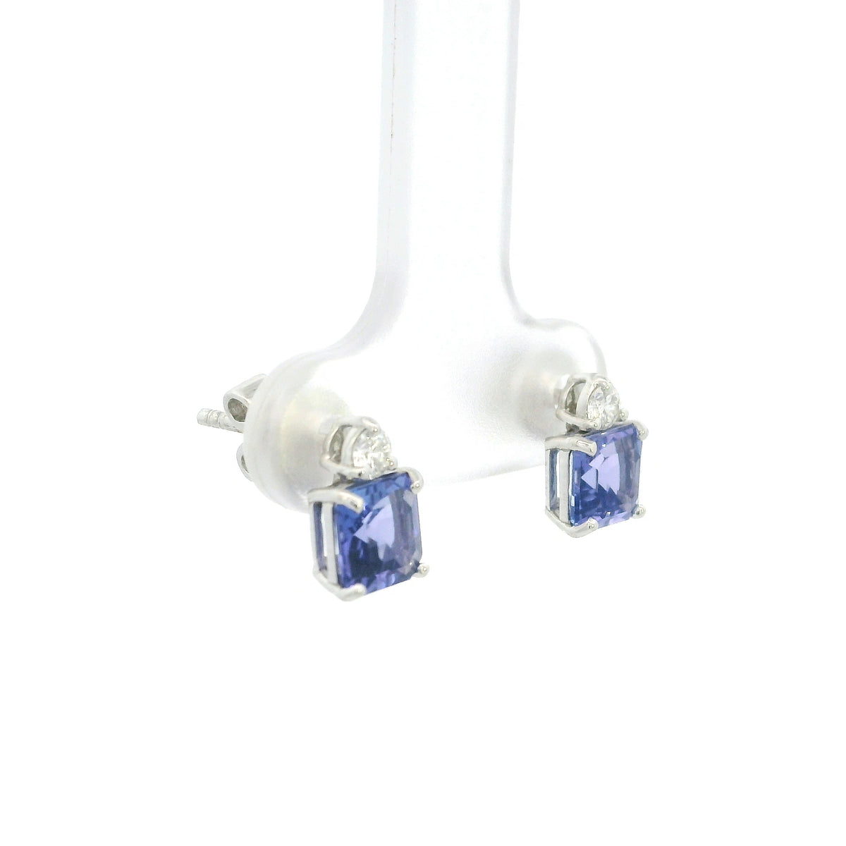 Pair of 18K white gold tanzanite and diamond stud earrings featuring square emerald-cut blue-violet tanzanites with a round diamond accent above each gemstone.