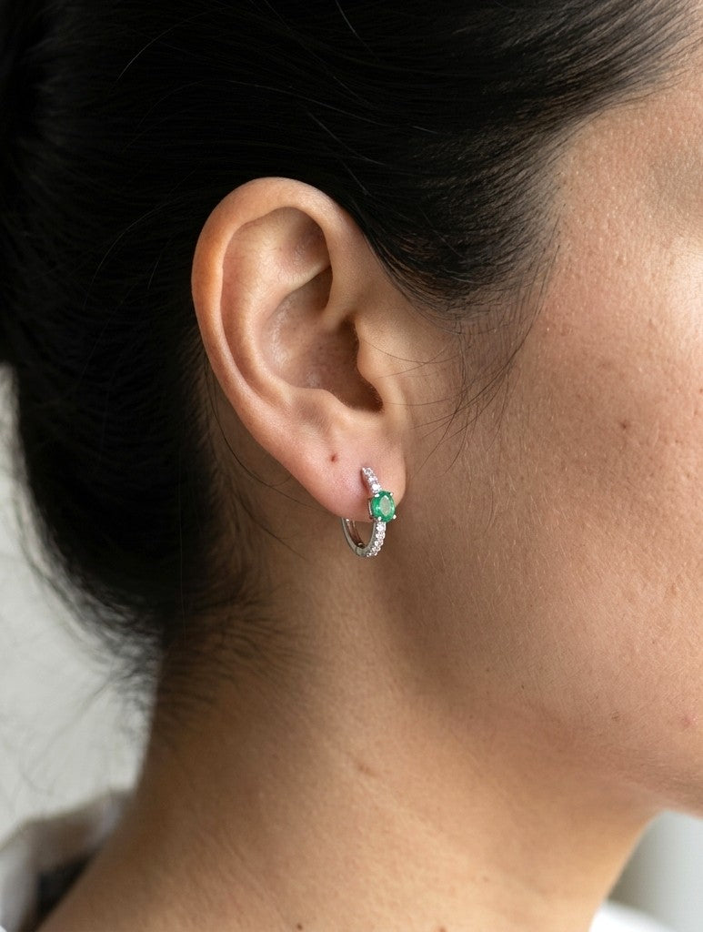 18K white gold emerald and diamond huggie hoop earrings featuring oval green emeralds and diamond-set hoops.