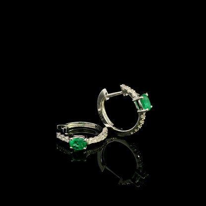 18K white gold emerald and diamond huggie hoop earrings featuring oval green emeralds and diamond-set hoops.