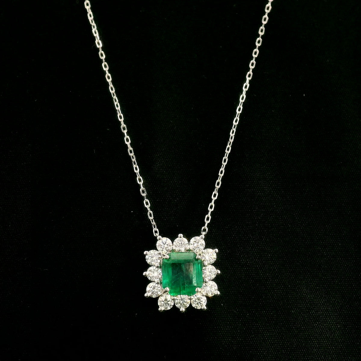 Emerald and diamond cluster halo necklace in 18K white gold featuring a 2.53 carat rectangular emerald surrounded by round diamonds totalling 1.46 carats, suspended from a fine white gold chain.