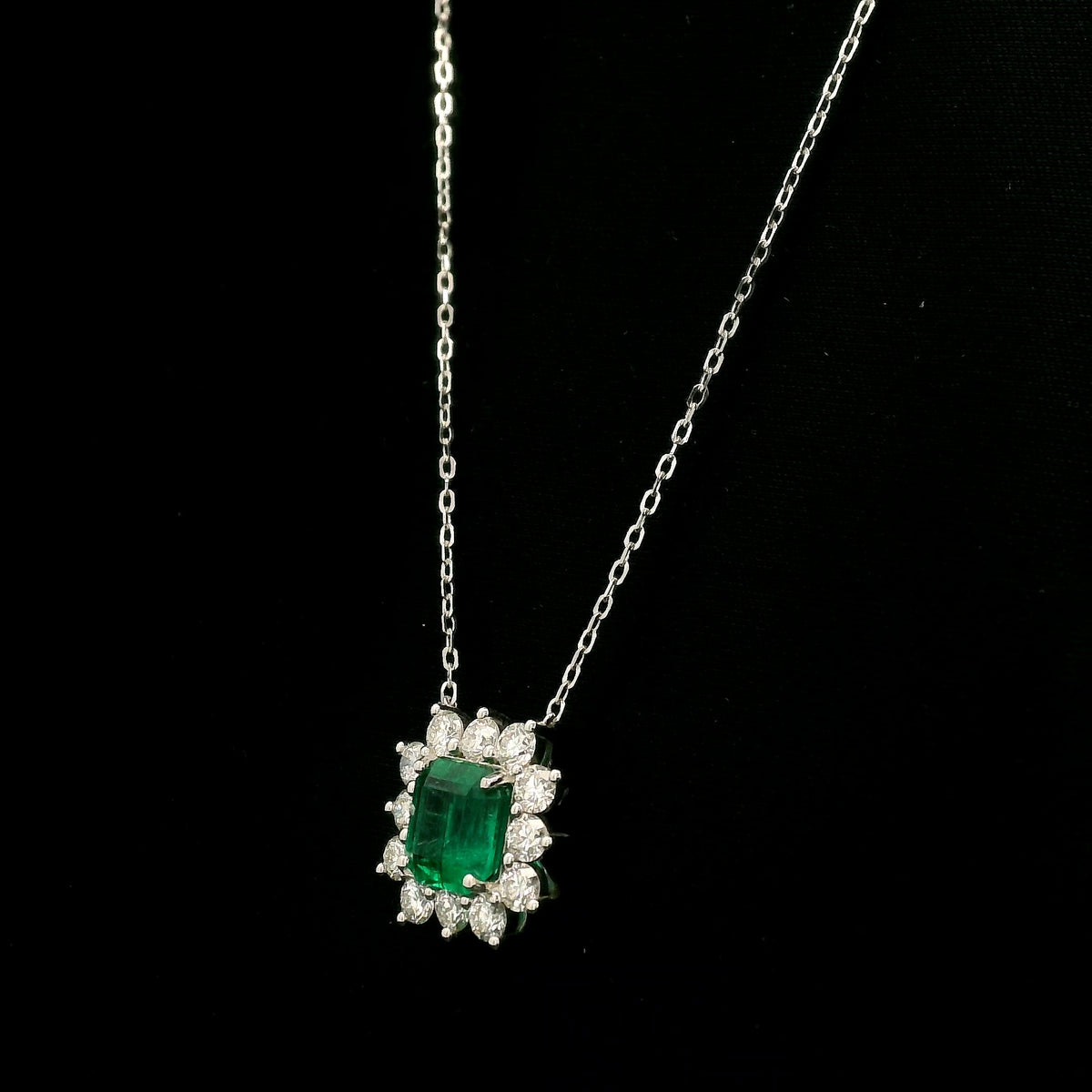 Emerald and diamond cluster halo necklace in 18K white gold featuring a 2.53 carat rectangular emerald surrounded by round diamonds totalling 1.46 carats, suspended from a fine white gold chain.