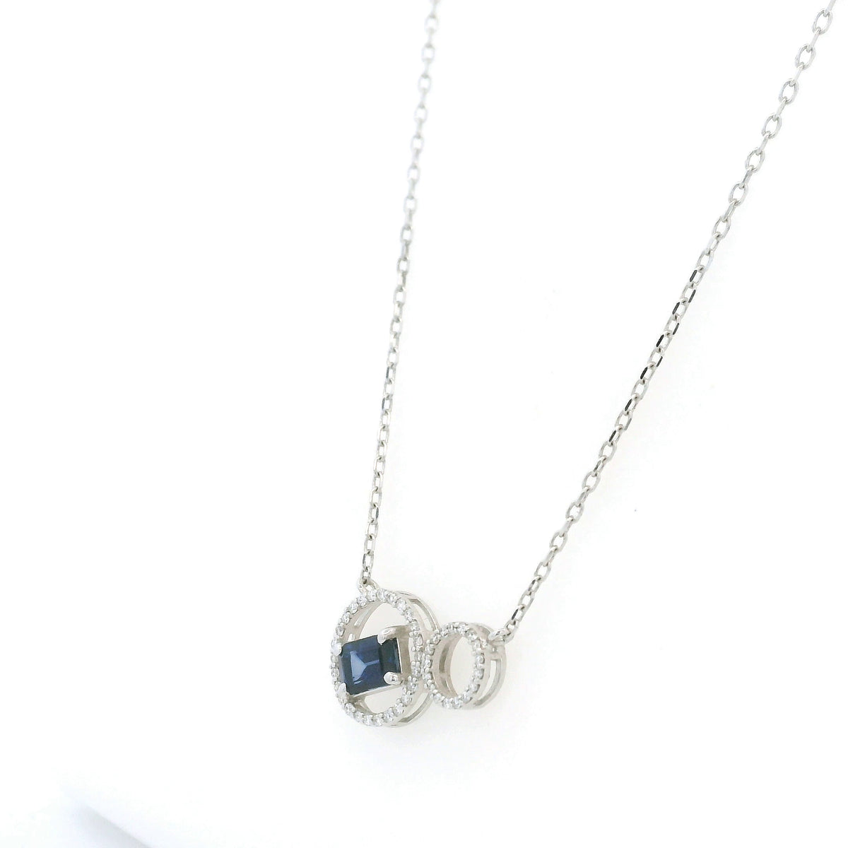 Blue emerald-cut sapphire and diamond necklace in 18K white gold, featuring an asymmetrical double-circle design with round diamond accents.