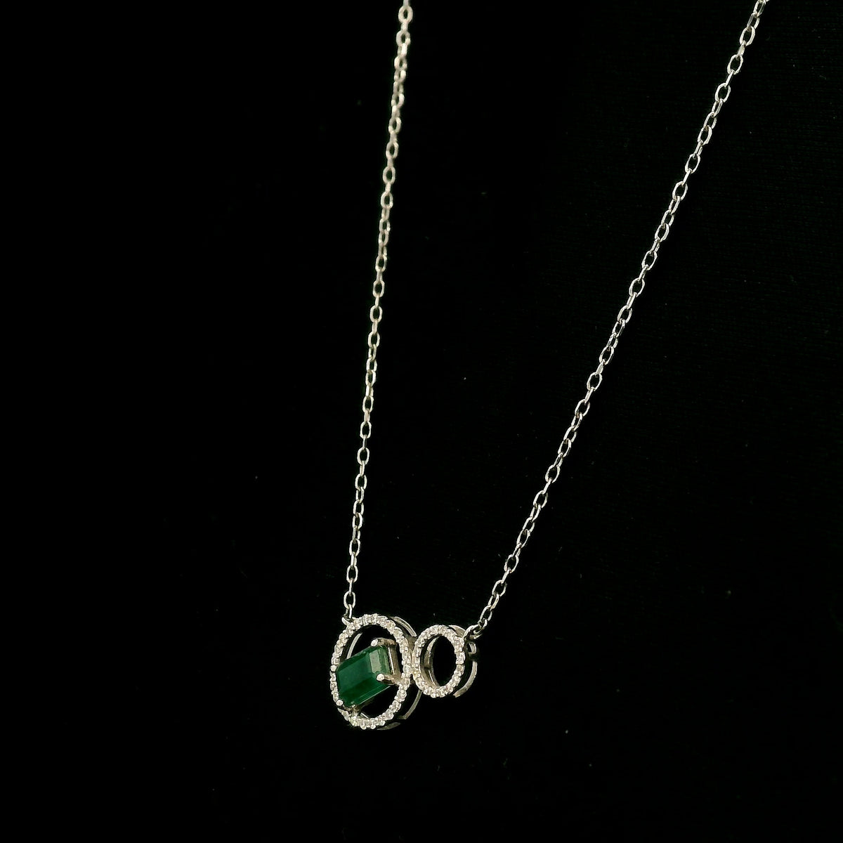 Emerald and diamond necklace in 18K white gold featuring a rectangular emerald-cut green gemstone inside a diamond circle, connected to a smaller diamond-set circle.