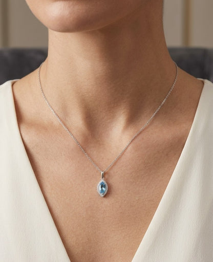 Blue topaz and diamond halo pendant in 18K white gold featuring a 0.70 carat oval blue topaz surrounded by 0.15 carat round diamonds in an elongated marquise-shaped setting, shown with a chain for display.