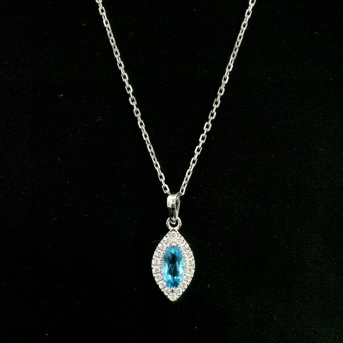 Blue topaz and diamond halo pendant in 18K white gold featuring a 0.70 carat oval blue topaz surrounded by 0.15 carat round diamonds in an elongated marquise-shaped setting, shown with a chain for display.