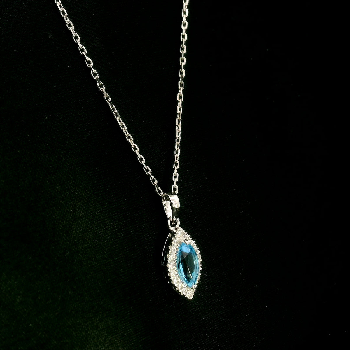 Blue topaz and diamond halo pendant in 18K white gold featuring a 0.70 carat oval blue topaz surrounded by 0.15 carat round diamonds in an elongated marquise-shaped setting, shown with a chain for display.