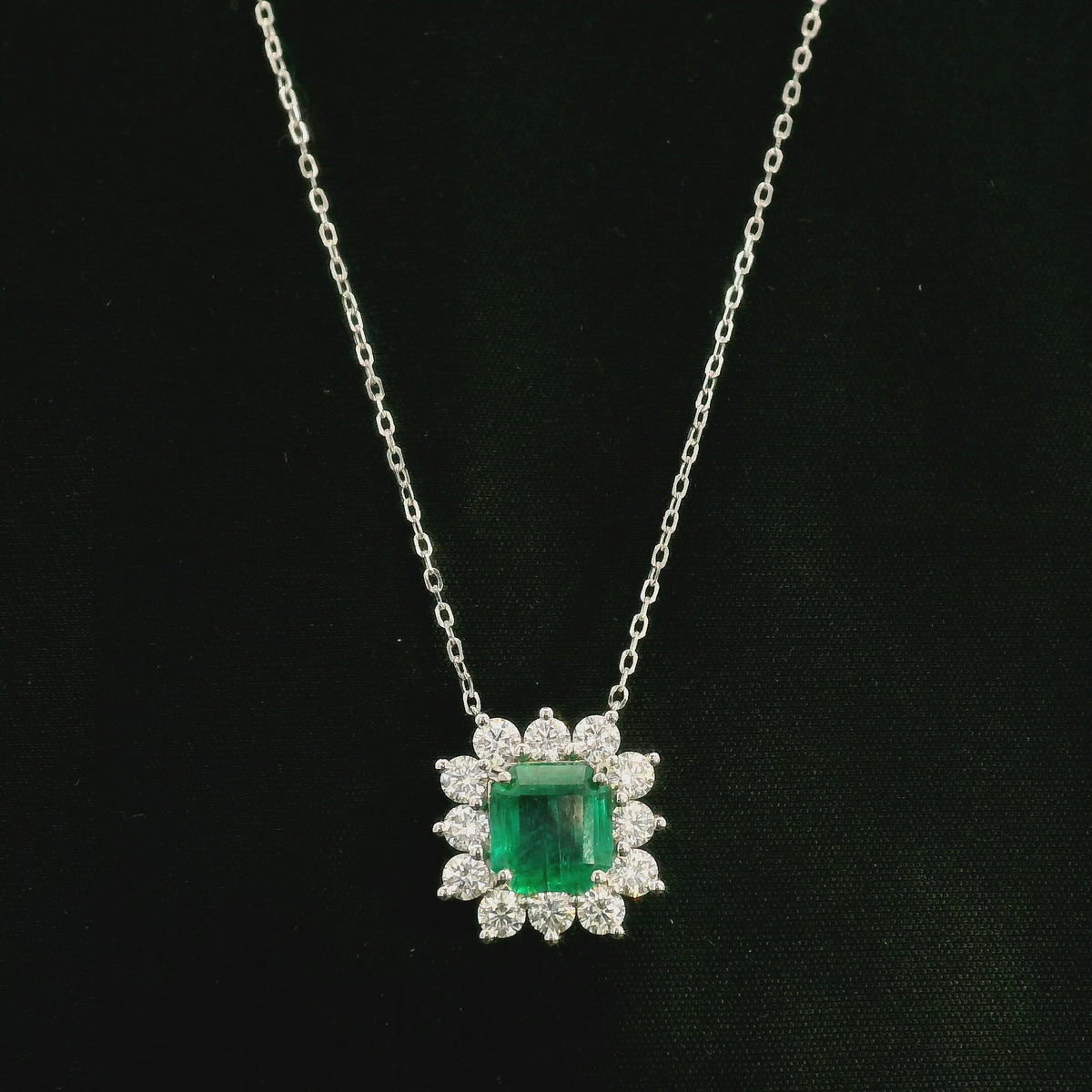 Emerald and diamond cluster halo necklace in 18K white gold featuring a 2.53 carat rectangular emerald surrounded by round diamonds totalling 1.46 carats, suspended from a fine white gold chain.