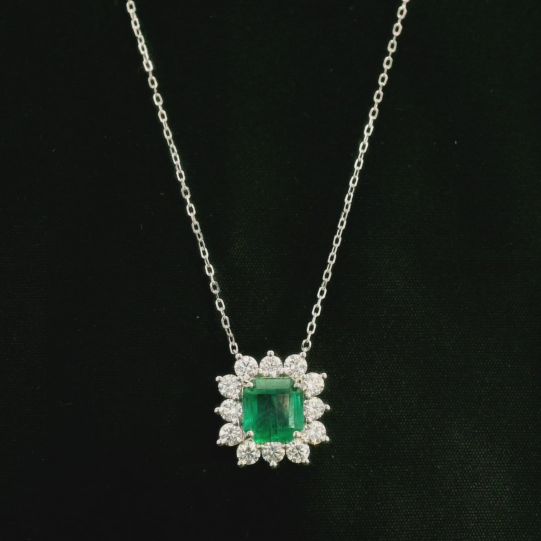 Emerald and diamond cluster halo necklace in 18K white gold featuring a 2.53 carat rectangular emerald surrounded by round diamonds totalling 1.46 carats, suspended from a fine white gold chain.