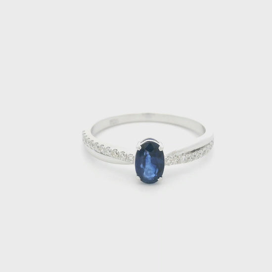 Oval blue sapphire ring with diamond-set shoulders in polished 18K white gold, featuring a four-prong centre gemstone setting.